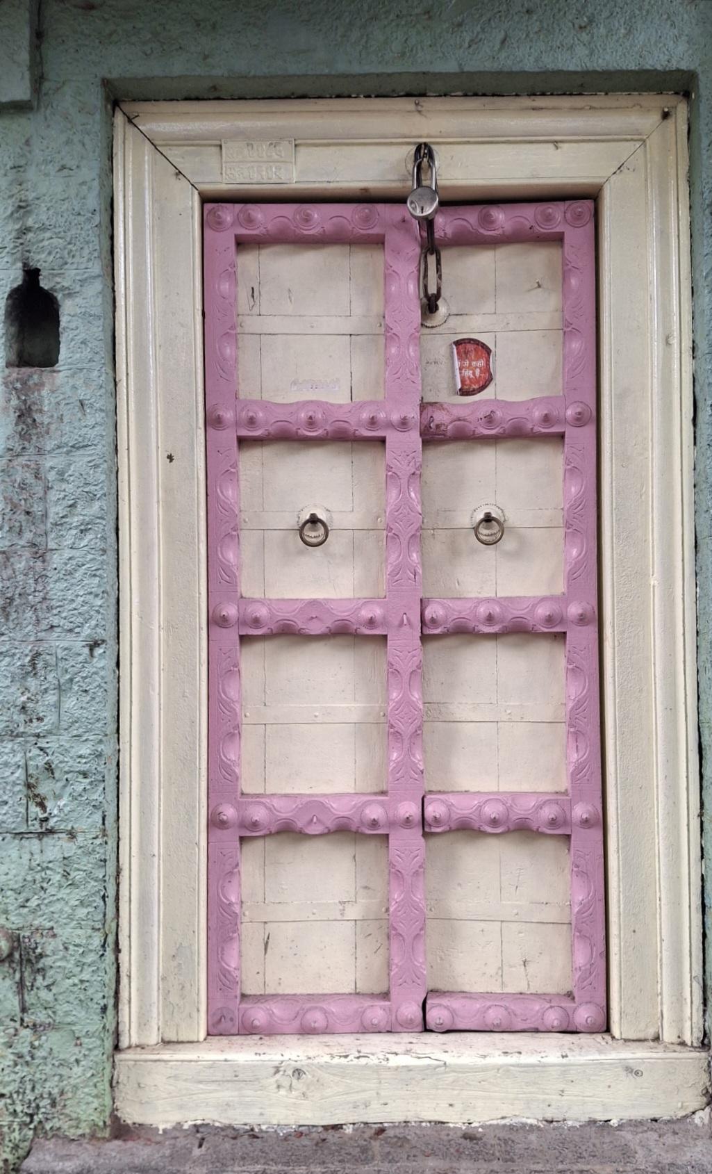 The back entrance doors of the house are painted in pink and white cream shades. The door features many interesting architectural elements. It has a distinctive design with the pink colored structure showcasing beautiful woodwork. The metallic round structures in the middle of the door are ring knockers and a devli can be spotted on the left of this door. (Source: CKA Archives)
