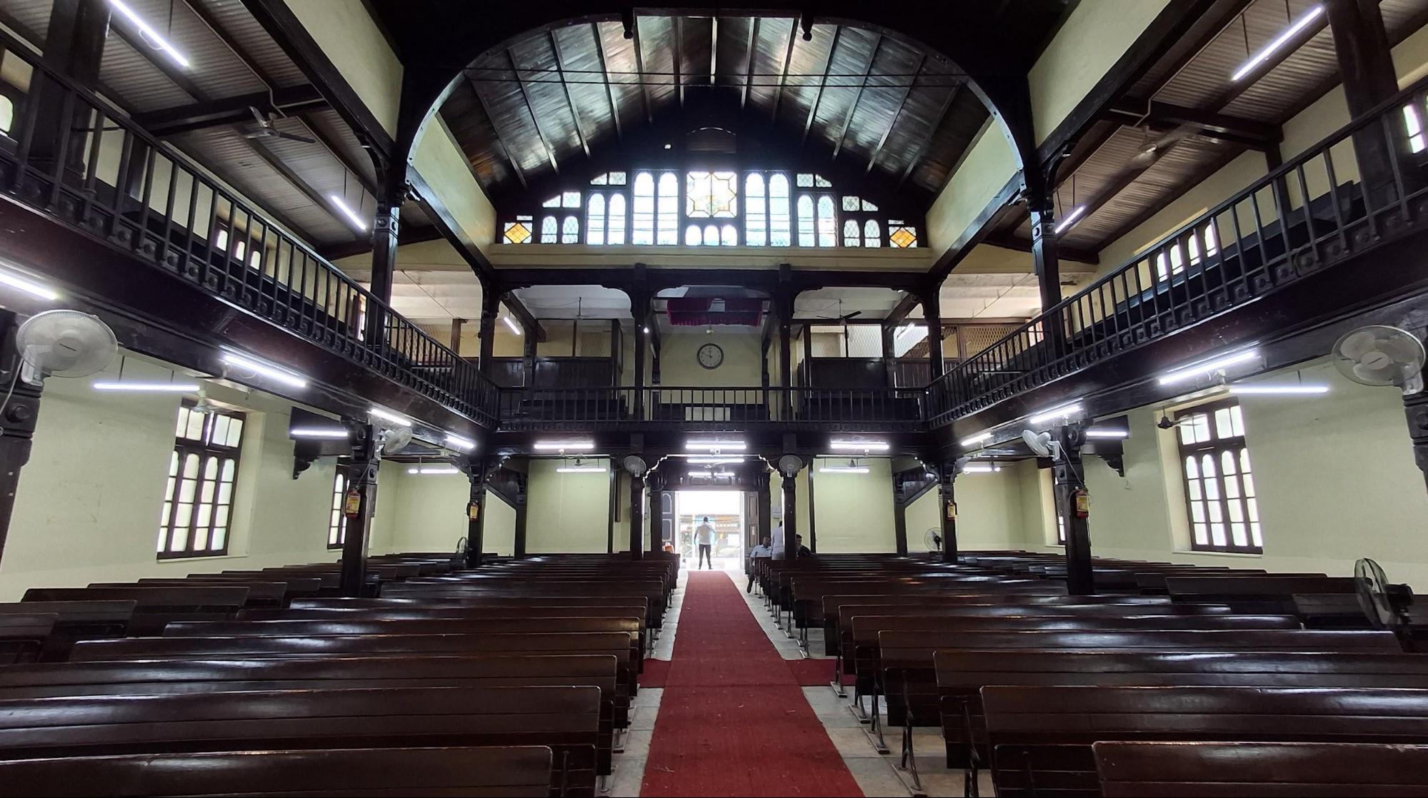 An interior view of Hume Memorial Church at Ahilyanagar showing the two-floor seating arrangement with wooden benches, teak woodwork, and central aisle with red carpet. (Source: CKA Archives)