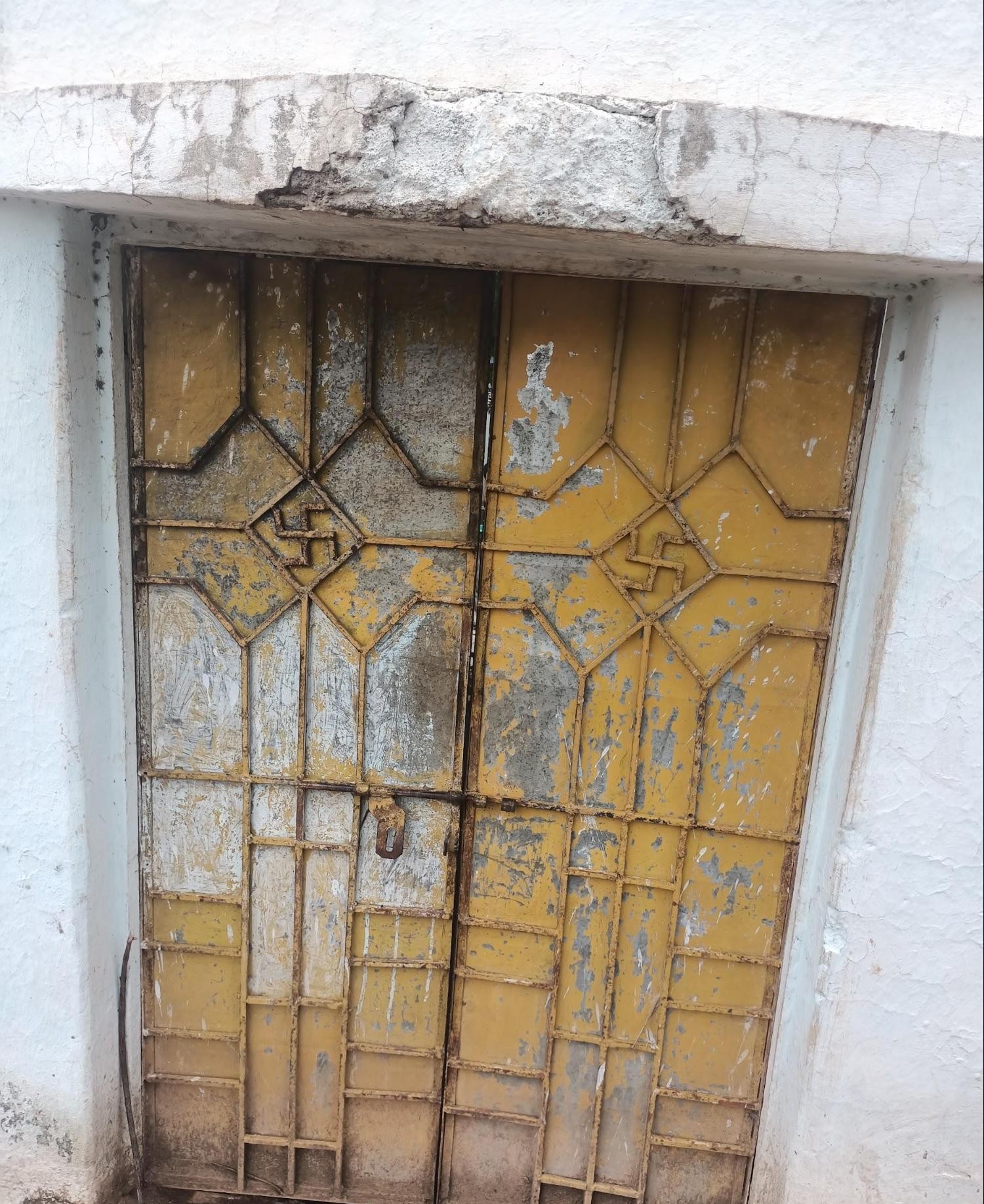 A double door with decorative metal overlay featuring swastika and geometric patterns, painted in ochre yellow with signs of aging.  (Source: CKA Archives)