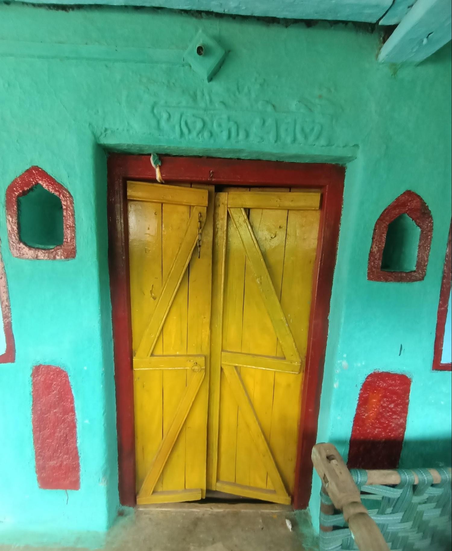 Bright yellow wooden door with Z-brace pattern, set against a turquoise wall and flanked by decorative red-painted niches on either side of the doorframe. It has some carved or embossed writing over it - could be the owner or family name. (Source: CKA Archives)