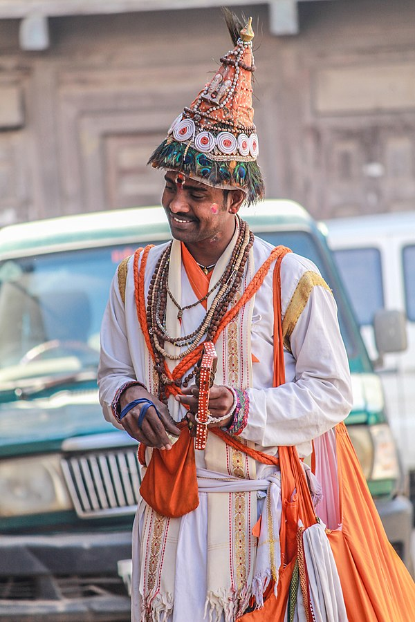 A Vasudev performer in traditional attire, wearing a cone-shaped hat adorned with peacock feathers, carrying a Tal while singing songs on Bhagwan Krishna.[3]