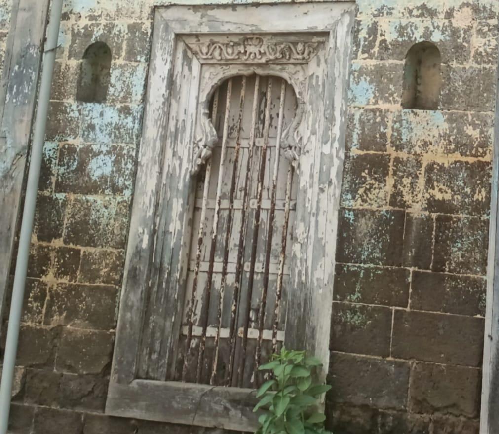 A weathered wooden door with vertical metal rods set within a stone frame. The top corners of the windows at the entrance of this wada have two sanas (curved elements that can be seen) carved onto them, presumably to put lanterns at night.  (Source: CKA Archives)